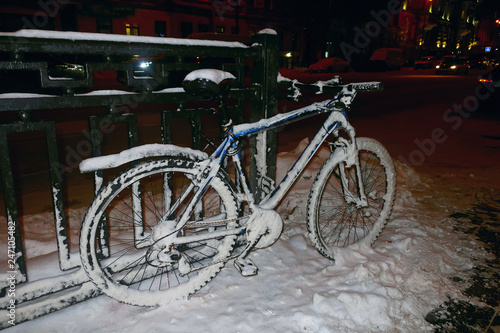 A snow-covered bike parked on a fence on a winter street.