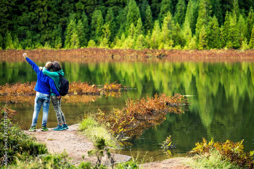 Young couple in colorful sweatshirts is standing in front of the volcanic lake on Azores Islands. Beautiful reflections on the background and beautiful green forest. Portugal.