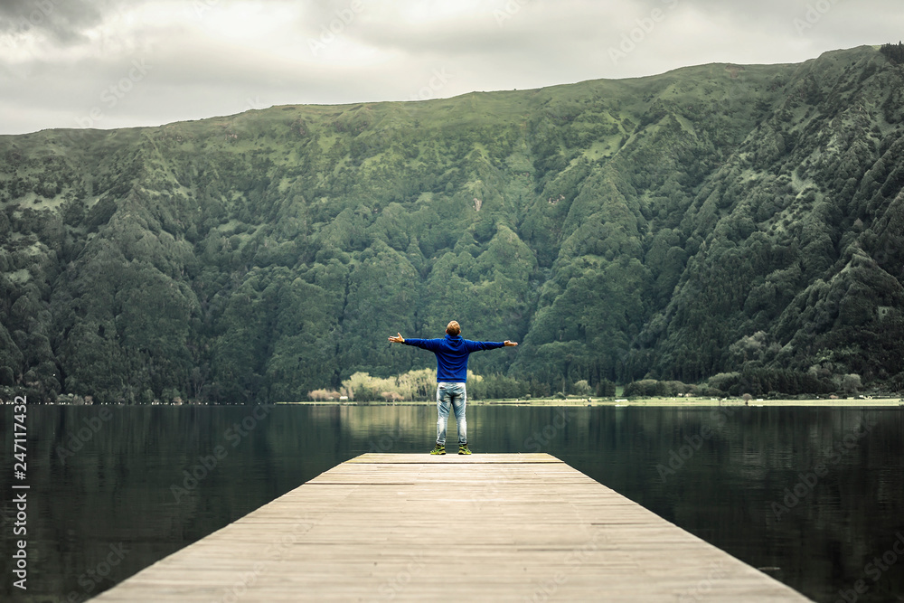 Young man in blue sweatshirt is standing on pier by the volcanic lake on Azores islands. Low angle view, soft focus. Mountain on background. Young man in blue sweatshirt is standing on pier by the volcanic lake on Azores islands. Low angle view, soft focus. Mountain on background.
