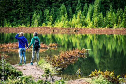 Young couple in colorful sweatshirts is standing in front of the volcanic lake on Azores Islands. Beautiful reflections on the background and beautiful green forest. Portugal.
