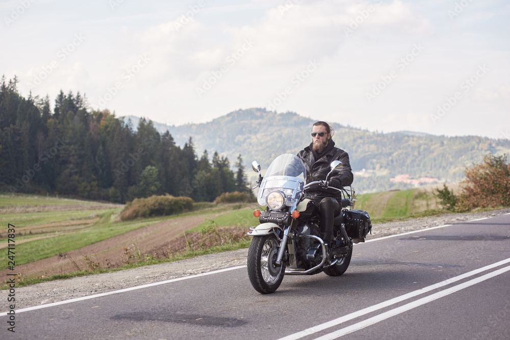 Handsome motorcyclist in black leather outfit riding cruiser bike on bright sunny summer day on background foggy distant green woody hills under morning sky.