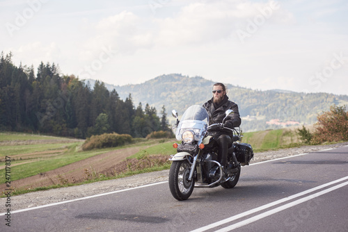 Handsome motorcyclist in black leather outfit riding cruiser bike on bright sunny summer day on background foggy distant green woody hills under morning sky.