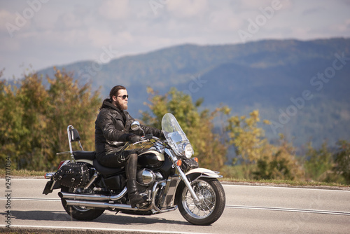 Side view of bearded long-haired motorcyclist in sunglasses and black leather clothing riding cruiser motorcycle along narrow asphalt path on sunny day