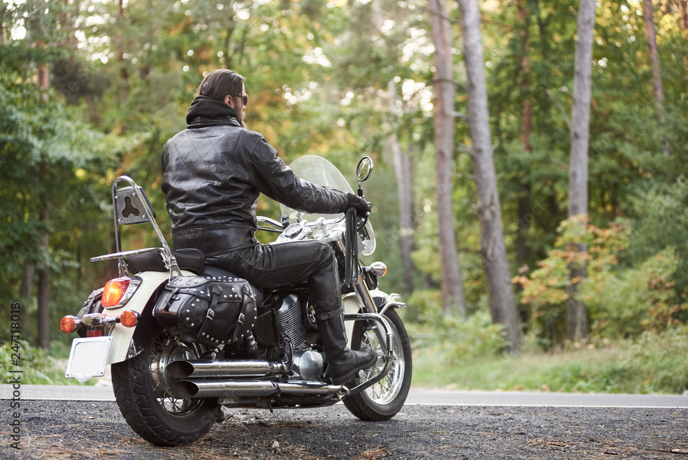 Back view of bearded biker in black leather clothing riding on cruiser motorbike on country roadside on background of empty straight asphalt road and green trees bokeh foliage.