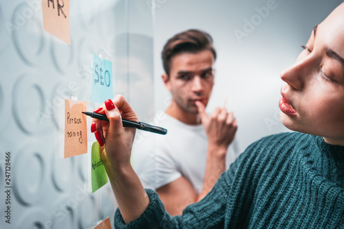 Intelligent coworkers people brainsorming new business plan posted on a sticky glass note wall. Team of HR manager having meeting standing behind stiky glass wall