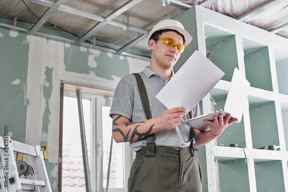 Room repair. Builder in helmet and glasses stands with a laptop and drawings in his hands on the background of the construction