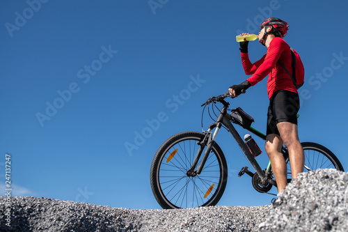 Cyclist resting and drinking isotonic drink. Backlight, sunny summer day. Extreme mountain bike sport athlete man riding outdoors lifestyle trail. © sutthinon602