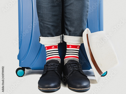 Stylish suitcase, men's legs, sunhat, bright socks with nautical theme and elegant shoes on a white, isolated background. Close-up. Concept of style, fashion, beauty and travel
