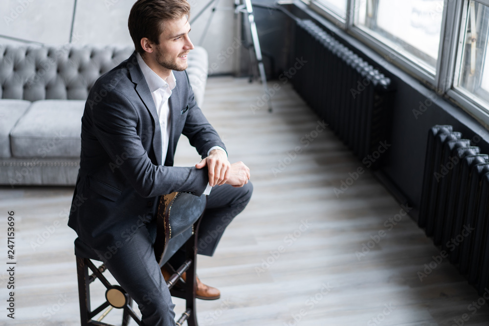 Portrait of confidence. Thoughtful young man in full suit looking away while sitting on the stool. Portrait of confidence. Thoughtful young man in full suit looking away while sitting on the stool.
