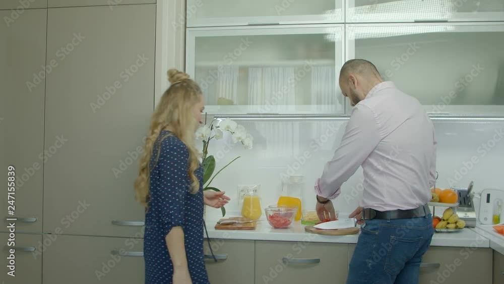 Caring man in smart casual clothes preparing morning breakfast in domestic kitchen. Husband slicing fresh tomatoes on cutting board, distracted by lovely housewife advices while cooking at home. Caring man in smart casual clothes preparing morning breakfast in domestic kitchen. Husband slicing fresh tomatoes on cutting board, distracted by lovely housewife advices while cooking at home.