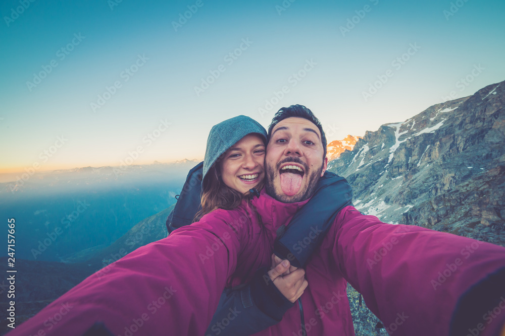 Couple of hikers taking selfie from top of the mountain with peaks and valley view on the background  after long trekking