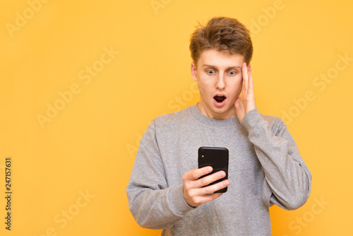 Portrait of a shocked guy standing with a smartphone in his hand on a yellow background, looking at the screen, took a hand on his head against the yellow background. Copyspace © bodnarphoto