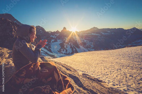 Girl sitting on a rock and covered with a blacket is watching Sunset From the top of the Mountain in a glacier area in the Alps with snow and blue sky