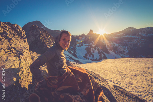 Girl sitting on a rock and covered with a blacket is watching Sunset From the top of the Mountain in a glacier area in the Alps with snow and blue sky photo