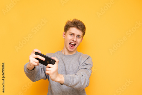 Portrait of a happy young man with a smartphone in his hands,smiling, looking into the camera and playing mobile games,isolated. Guy is a gamer with a smartphone in his hands on a yellow background