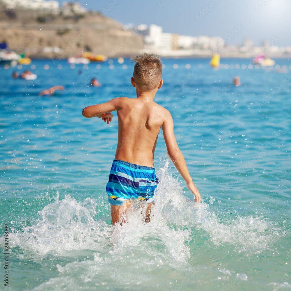 Cute European boy in a striped swimming shorts is running in to the sea. He is enjoying his summer holidays. Back view.