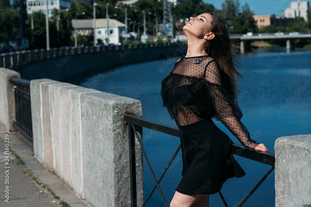 beautiful brunette stands on the bridge, near the river, in the city. a girl in a black dress puts a face to the sun and the wind that blows on her