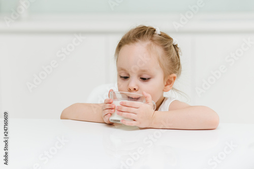 Little smart caucasian girl drinks milk from a glass cup in the kitchen and has fun, healthy eating concept for children.