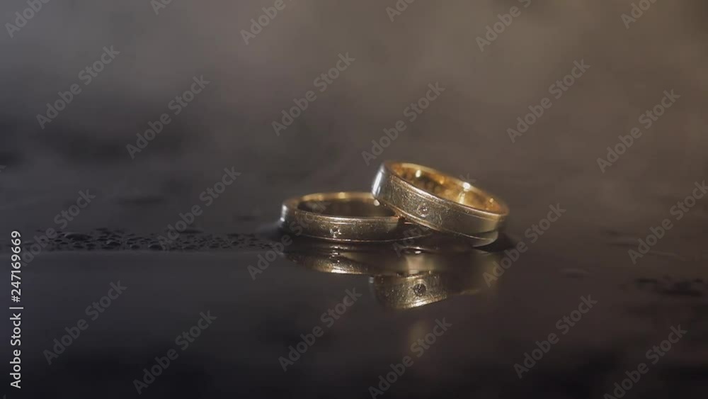 Wedding rings lying on dark surface with reflection and water. Shining with light close up macro. Smoke. Slow motion Wedding rings lying on dark surface with reflection and water. Shining with light close up macro. Smoke. Slow motion