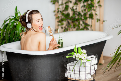 Young relaxed woman listening to the music and drinking smoothie while lying in the retro bathtub at the beautiful bathroom with green plants