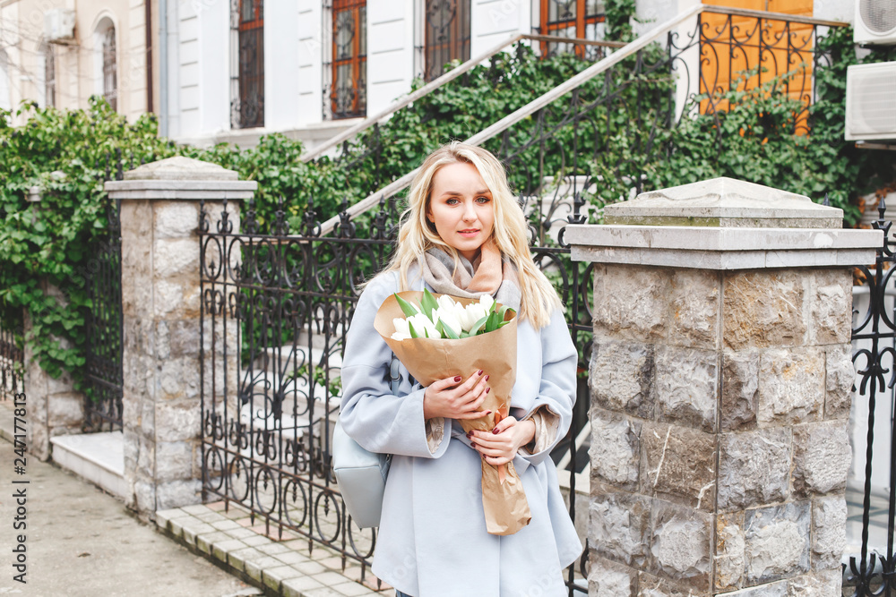 Young beautiful blonde woman in blue coat with bouquet of white tulips flowers walking outdoors on the city street in spring.