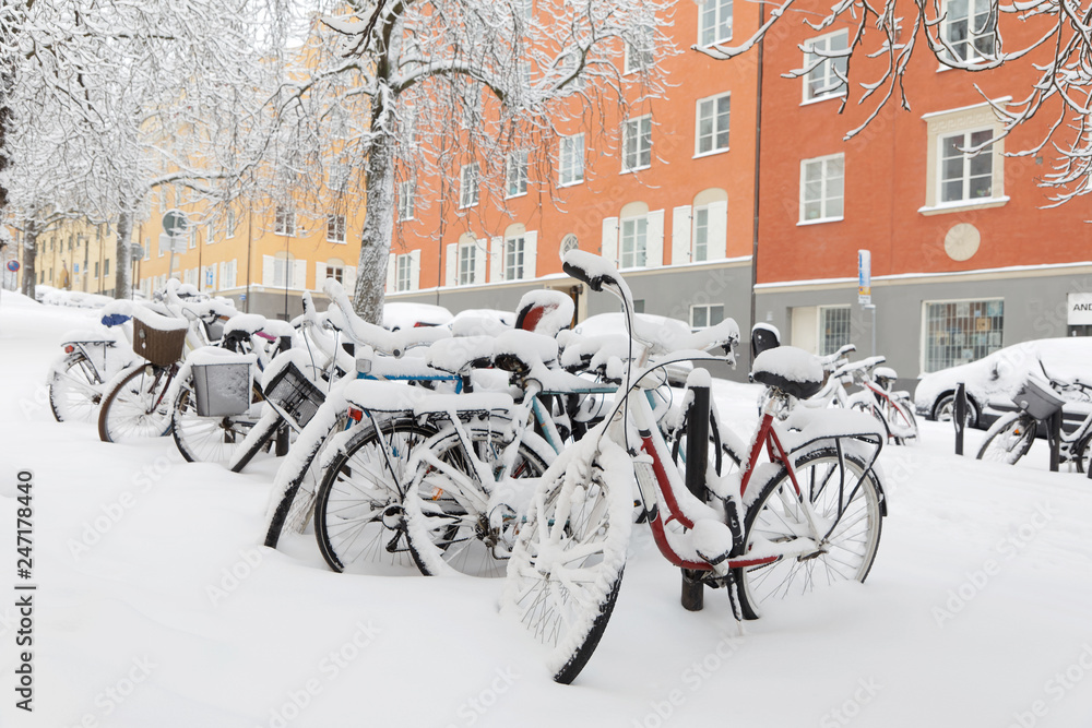 Snow chaos in the city, parked bicycles covered with snow, trees and buildings in the background