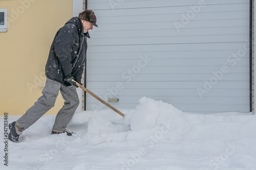 A man in a black jacket and light-colored gray pants and a brown hat cleans snow with a wooden shovel in winter