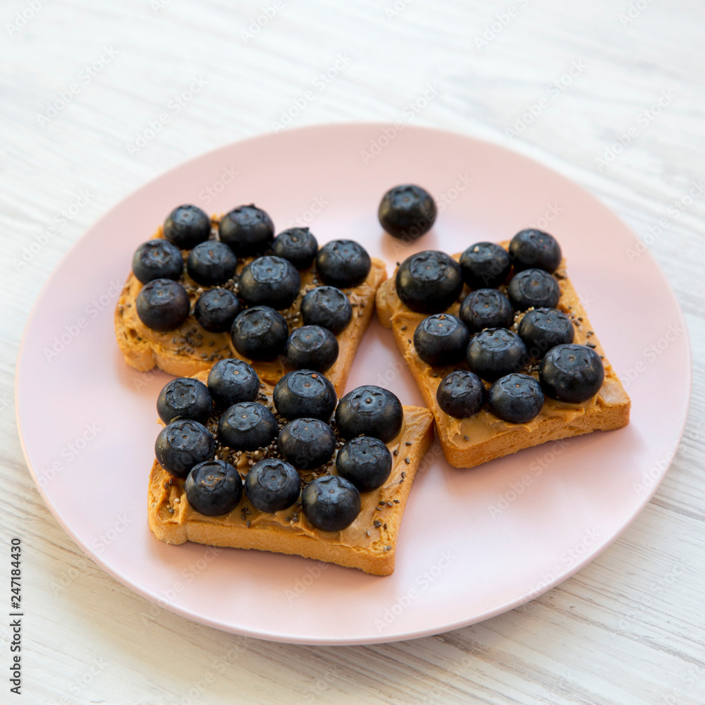 Fototapeta premium Vegetarian toasts with peanut butter, blueberries and chia seeds on a pink plate over white wooden surface, side view. Healthy eating and dieting.