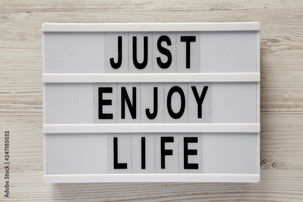 Modern board with text 'Just enjoy life' on a white wooden background, top view. Modern board with text 'Just enjoy life' on a white wooden background, top view.