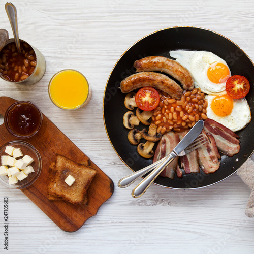 English breakfast in a frying pan with fried eggs, bacon, sausages, beans and toasts on white wooden surface, overhead view. Flat lay. From above.