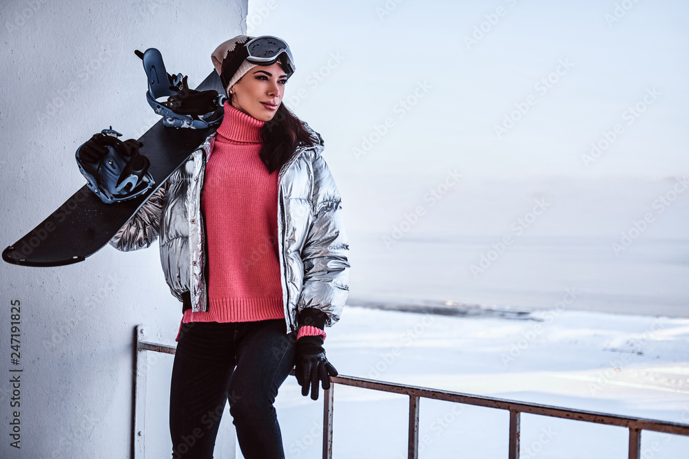 A beautiful woman wearing stylish warm clothes and goggles posing with snowboard, looking away while leaning on a railing near a snowy beach