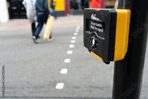 Close up of yellow metal crosswalk button for pedestrian crossing signal in London, UK for traffic rules. Two peoples crossing street by using pedestrian cross button.