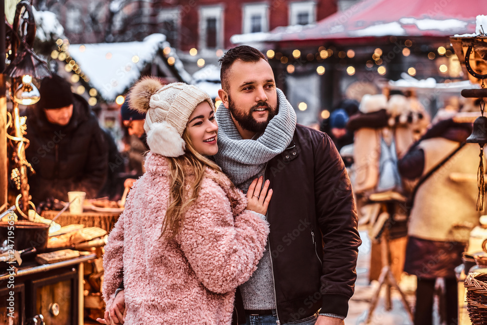 A happy attractive couple wearing warm clothes standing together near a shop counter of a winter fair at a Christmas time. A happy attractive couple wearing warm clothes standing together near a shop counter of a winter fair at a Christmas time.