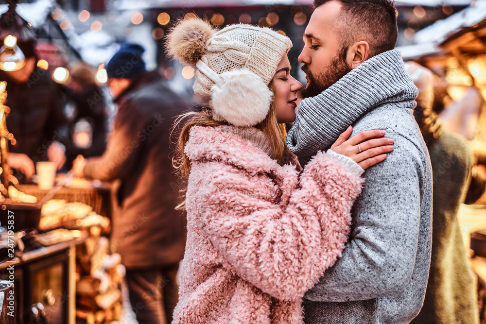 An attractive couple in love, a stylish couple wearing warm clothes cuddling together and looking each other at the winter fair at a Christmas time. An attractive couple in love, a stylish couple wearing warm clothes cuddling together and looking each other at the winter fair at a Christmas time.