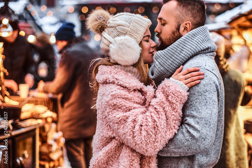 An attractive couple in love  a stylish couple wearing warm clothes cuddling together and looking each other at the winter fair at a Christmas time.