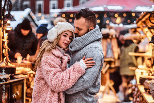 An attractive couple in love, a handsome man and charming girl cuddling and enjoying spending time together while standing at the winter fair at a Christmas time