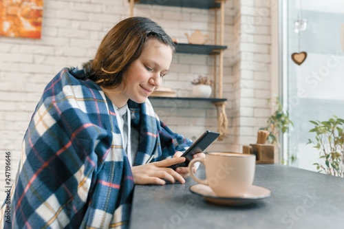 Autumn winter portrait of smiling teen girl with mobile phone and cup of coffee in coffee shop  girl covered with woolen plaid blanket