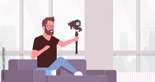 man blogger broadcasting live stream shooting selfie video guy in front of DSRL camera recording himself using gimbal stabilizer social media concept living room interior horizontal