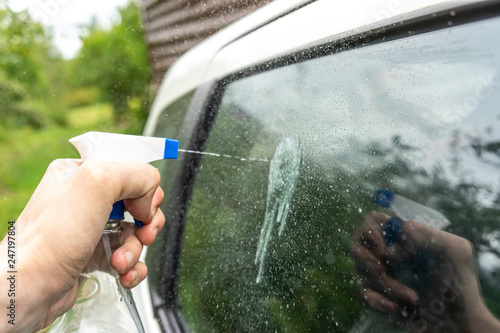 A hand holding a plastic spray bottle splashes onto a car's glass.