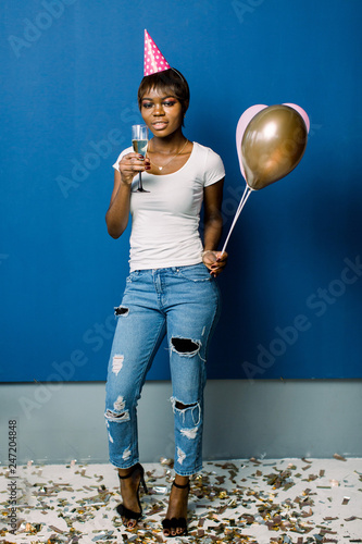 Full length portrait of a cheerful young african woman holding bunch of air balloons and champagne isolated over blue background. Birthday party, celebration concept.