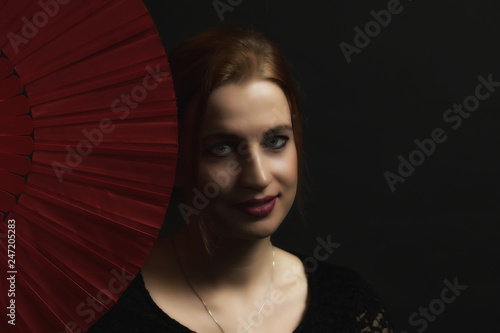 Low Key portrait of seductive young woman holding red fan. All on the dark background.