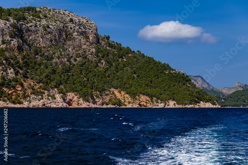 sunny day, sea view to the coast; view of the white waves left by the motorboat; on the shore there are large mountains partially overgrown with trees