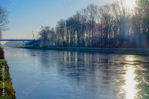 Beautiful early winter landscape with a frozen river or canal, treelined riverside and grass at sunrise creating a tranquile and quiet scenic nature background © Bjorn B
