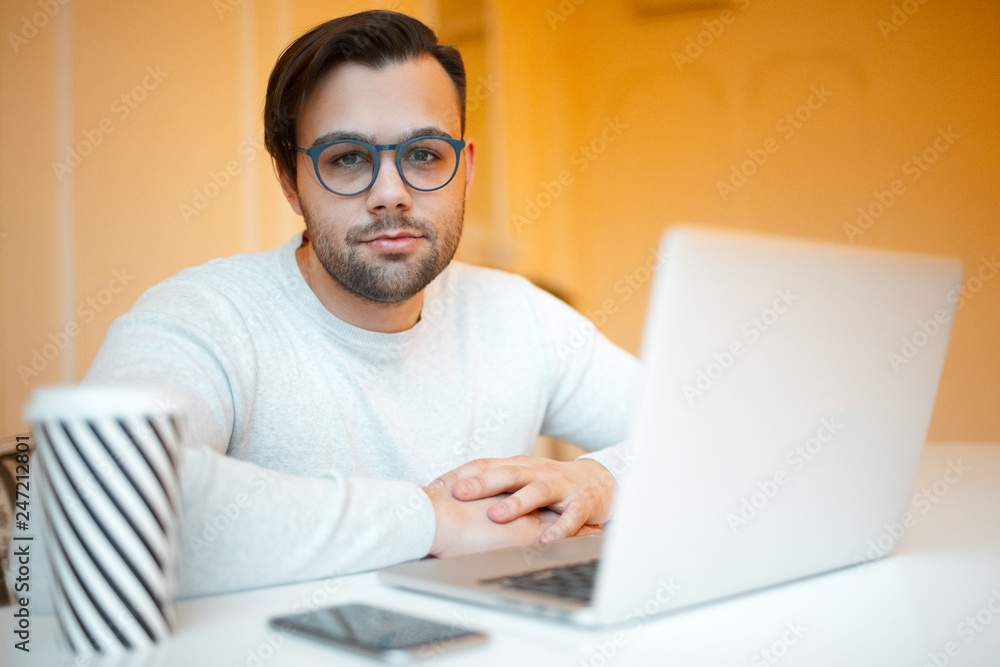 Young businessman with laptop, smartphone and coffee on white table. Wearing blue glasses and sweater, over yellow background.