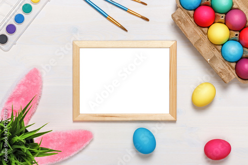 A frame, bunny ears, a flower pot with artificial green grass, colorful eggs in a cardboard box, paints and brushes. Easter concept. Copy space