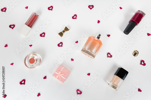 Cosmetic flat lay - perfume, nail polish, lipstick, small gift box, golden accessories on white background with tiny red hearts among them. Cosmetic sale, promotion, advertisement. 