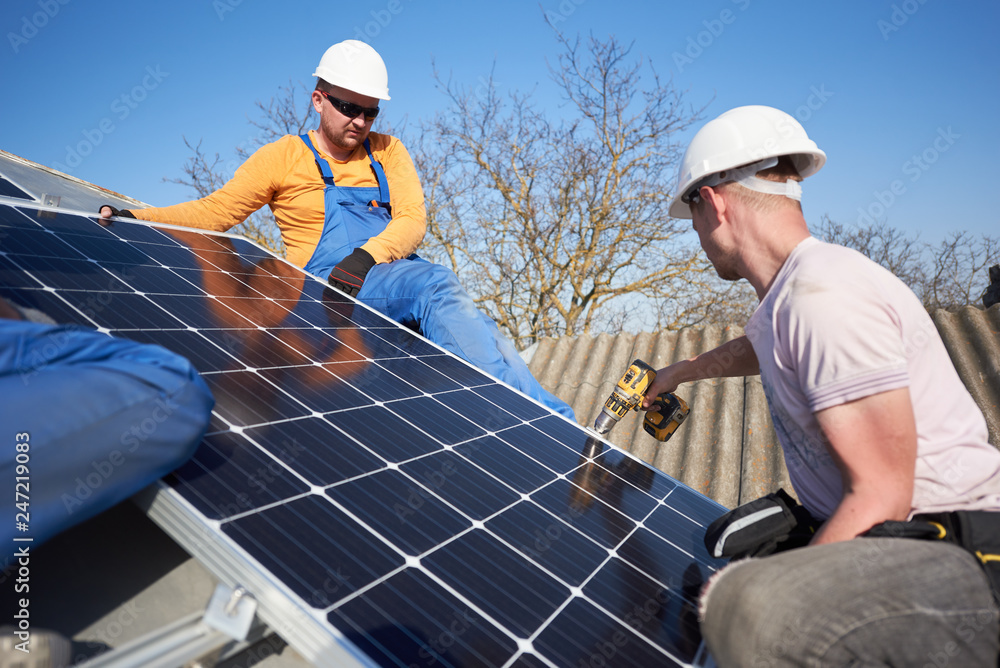Male team workers installing stand-alone solar photovoltaic panel system using screwdriver. Group of electricians mounting solar module on roof of modern house. Alternative energy ecological concept.