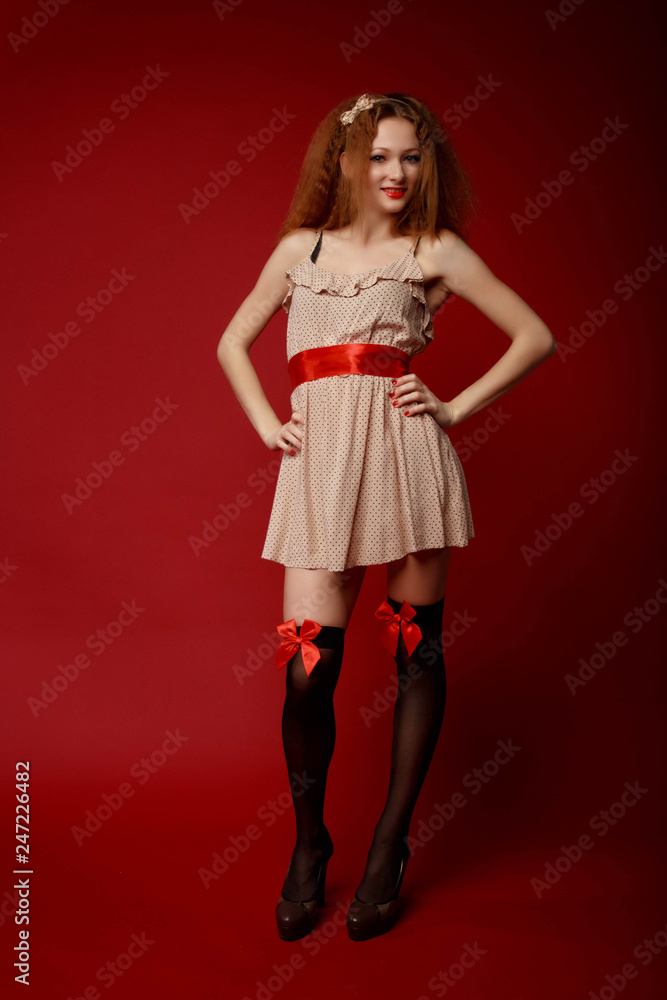 Pretty young woman in short dress, red belt,black stockings with ribbons and high heels poses with her hands on the hips against the red background. Pin-up full-growth retro-style portrait Pretty young woman in short dress, red belt,black stockings with ribbons and high heels poses with her hands on the hips against the red background. Pin-up full-growth retro-style portrait