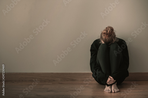 Sad teenager girl sitting curled up on the floor, copy space on empty wall