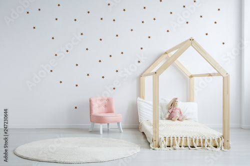 Pastel pink armchair next to wooden house shape bed with toy and blanket, copy space and golden stars on empty white wall, round white carpet on floor